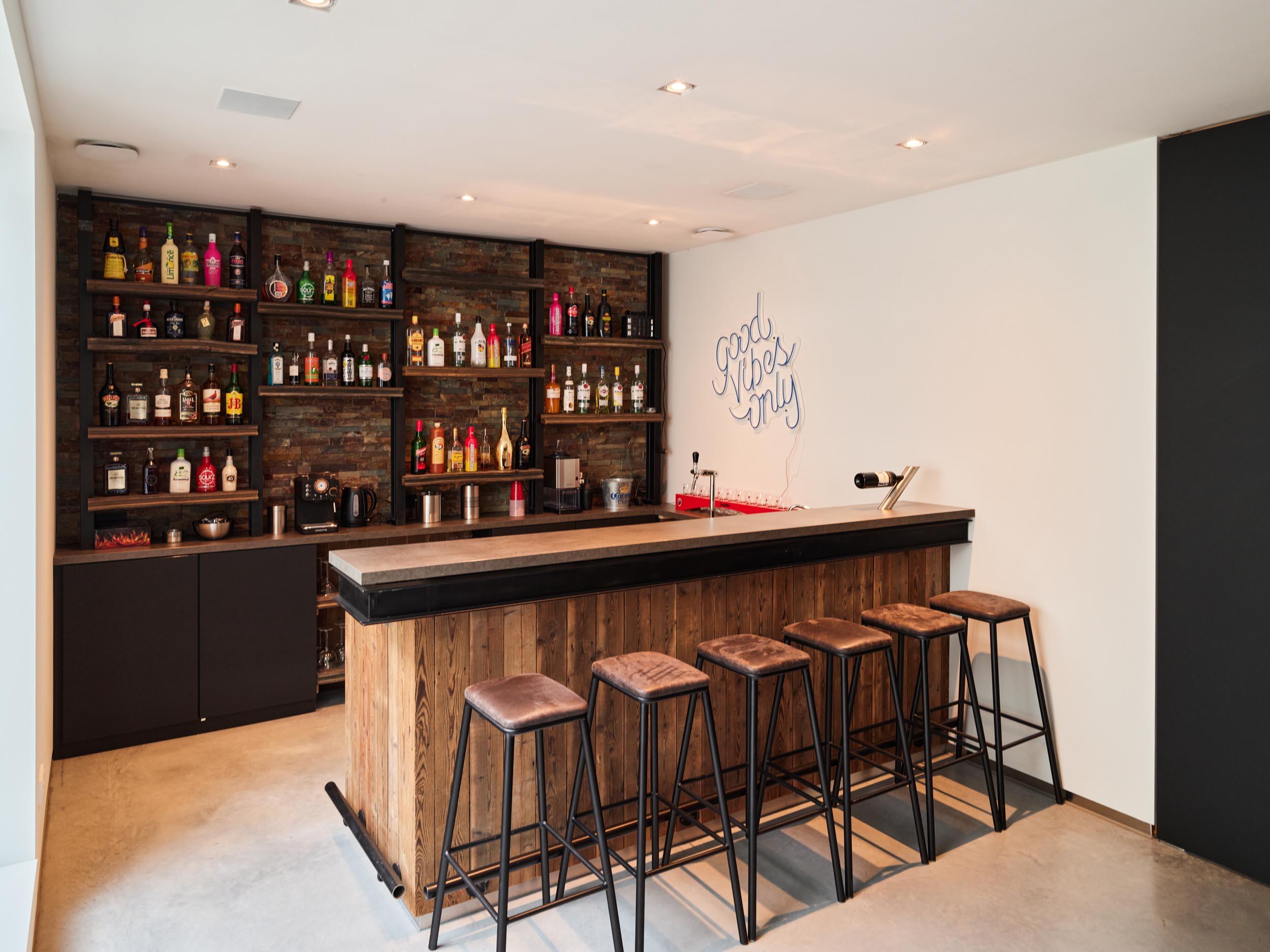 Stijlvolle huisbar met een naadloos akoestisch plafond, houten toog, barkrukken, een achterwand vol drankflessen en een neonbord met de tekst “good vibes only”